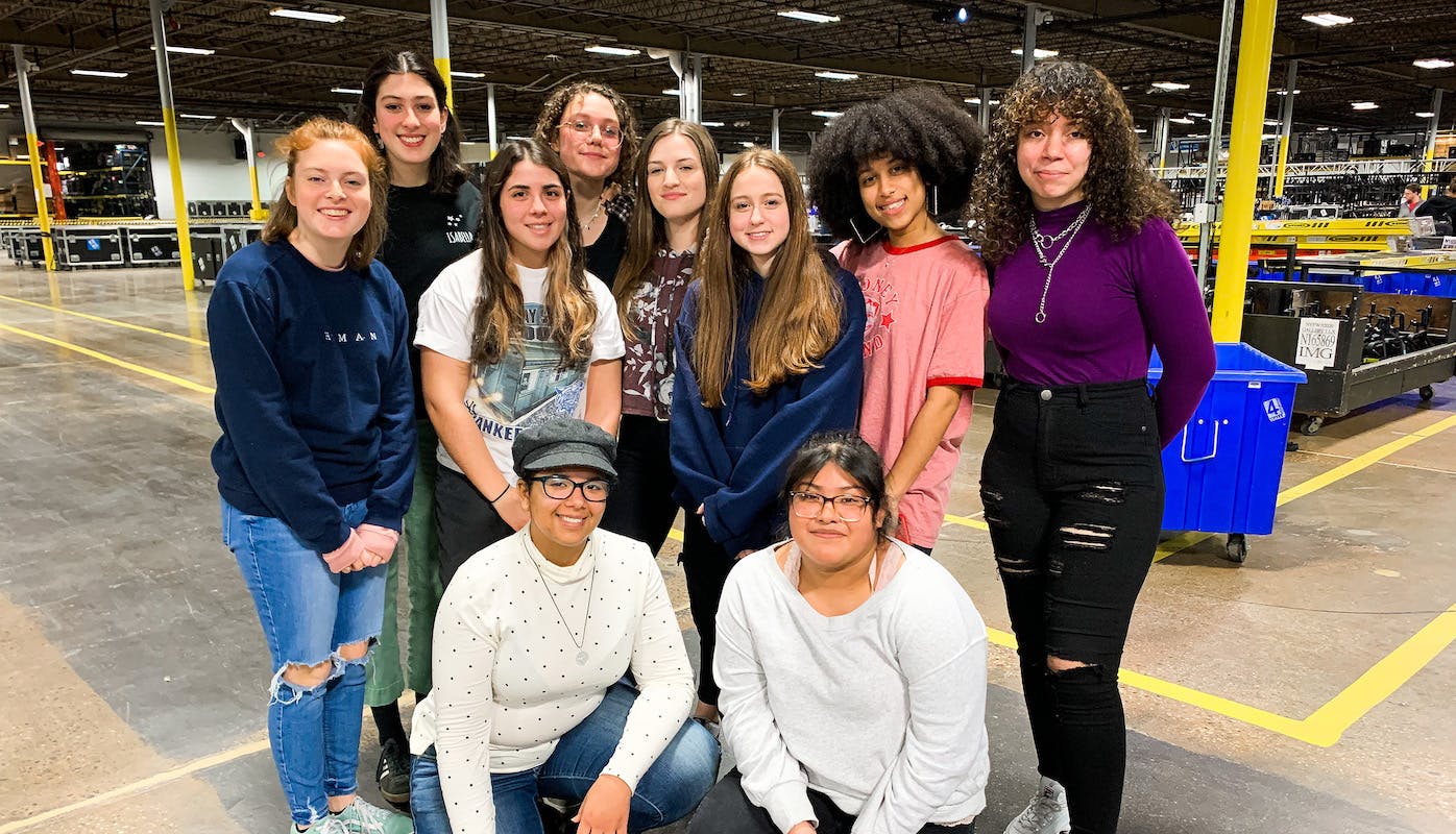 10 Open Stage Project students pose for a photo after a tour of 4Wall Entertainment. Their backs face a large warehouse with organized lighting equipment and storage boxes.