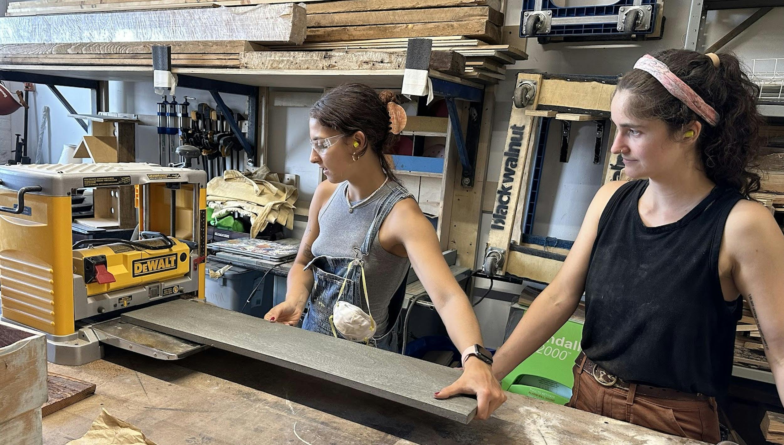 A photo taken in a carpentry studio with shelves of lumber and equipment in the background. A young woman, wearing clear safety-glasses and ear protection, uses a planer on a long piece of wood under the supervision of another woman.