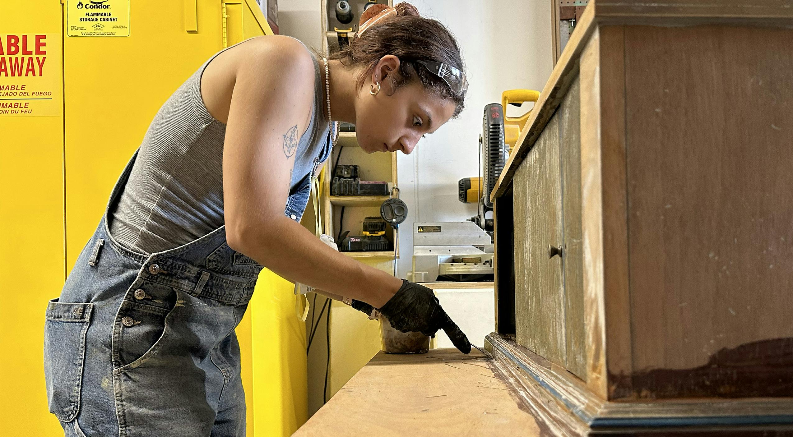 A young woman, wearing a gray top and denim overalls with gloves and safety glasses, works on a wooden desk in a scene shop.