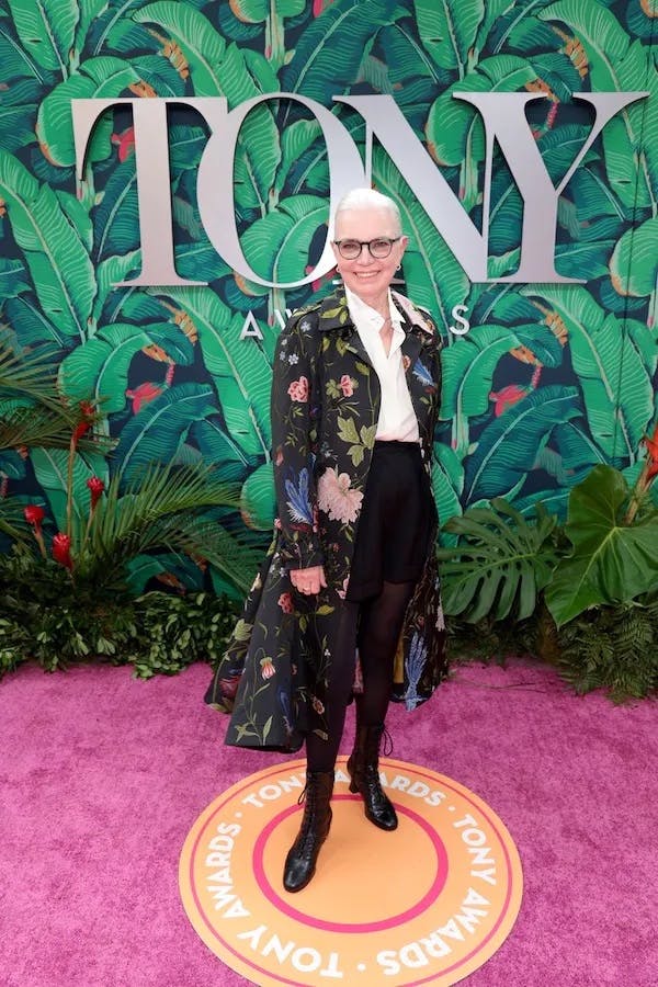 Photo of a woman in a stylish suit with a long floral coat, posing on the pink carpet of the Tony Awards.