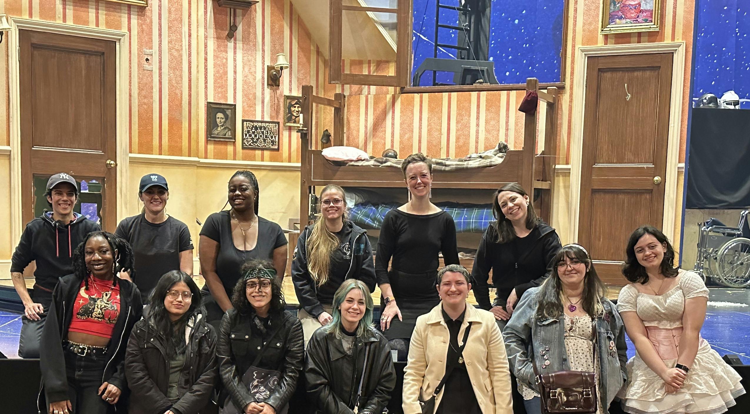 Students from Open Stage Project pose with female and non-binary crew members in front of the set of Peter Pan Goes Wrong on Broadway after a performance.