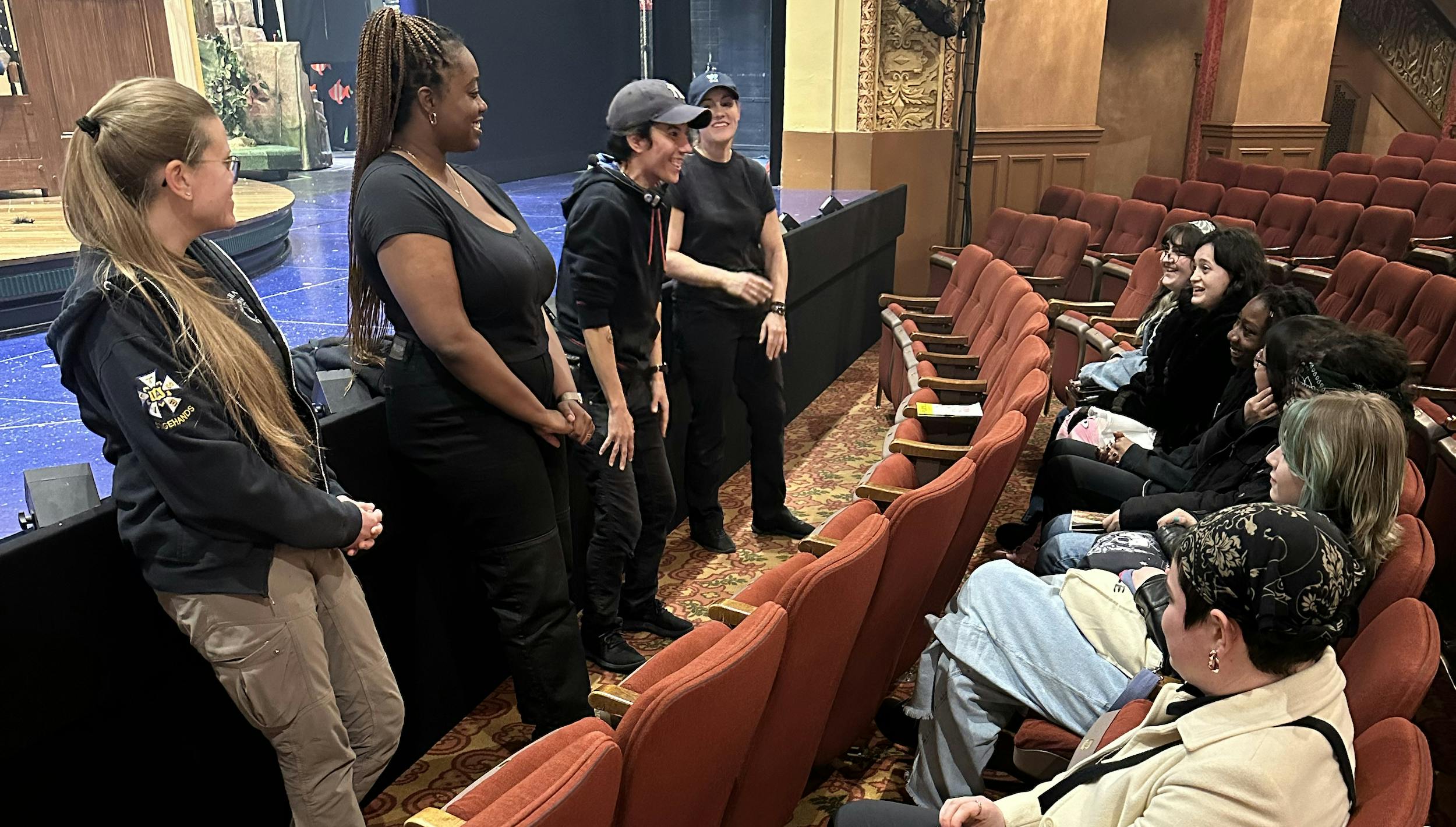 Engaged students sit in a row of theater seats near the front of the stage and talk to a group of female and non-binary crew members who stand in a line on the floor in front of the stage.